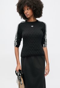 Čierna textúrovaná mikina s tromi bielymi pruhmi na každom rukáve a malým bielym logom na hrudi. Dopĺňa ju čierna sukňa a kabelka.