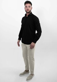 Man staat tegen een witte achtergrond met een zwart overhemd met knopen, beige broek en beige instapschoenen, kijkt recht vooruit.