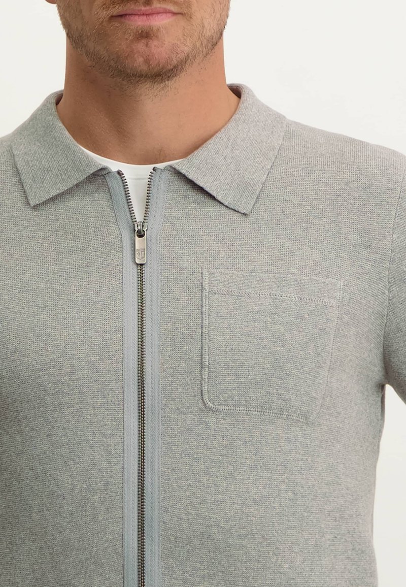 Grijze ritssluiting cardigan gemaakt van gestructureerde brei stof. Bevat een kraag en een borstzak. De ritssluiting loopt over de hele voorkant.