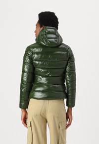 Glanzende olijfgroene pufferjack met horizontale quilting, een aansluitende capuchon en elastische manchetten, gecombineerd met beige cargobroeken.