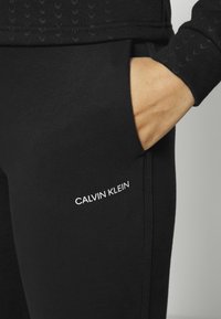 Person's hand resting inside the pocket of black Calvin Klein sweatpants with the brand name printed in white on the thigh.