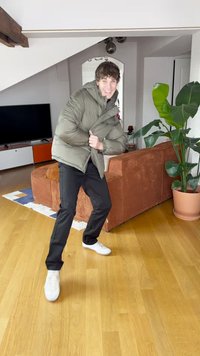 Olivengrøn polstret jakke, sorte bukser og hvide sneakers. Indendørs miljø med trægulv og en brun manchester-sofa.