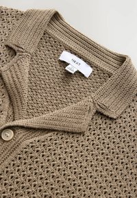 Beige strikket cardigan med krave, der har et åbent vævemønster og en knaplukning. Etiket vist ved kraven.