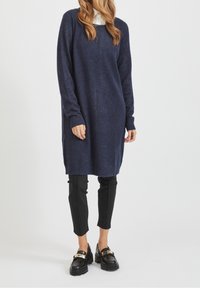 Femme portant un long pull en tricot bleu marine, un pantalon slim noir et des mocassins noirs épais, debout devant un fond blanc uni.