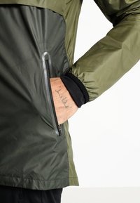 Main avec un tatouage partiellement à l'intérieur de la poche zippée d'une veste bicolore vert olive avec un poignet intérieur noir.