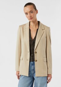 Beiger gestreifter Blazer mit Reverskragen, zwei Knöpfen und Vordertaschen, getragen über einem dunklen Oberteil und hellblauen Jeans.