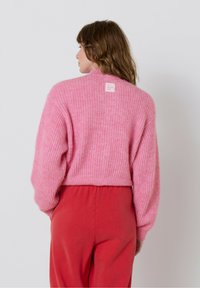Ružový rebrovaný sveter s kratším strihom, so sťahovacími dlhými rukávmi a malou značkou na chrbte. Spárované s červenými nohavicami na elastický pás.