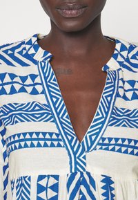 Femme africaine portant un vêtement bleu et blanc à motif avec un col en V et un tatouage léger visible sur sa poitrine.