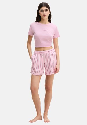 Frau steht barfuß, trägt ein pinkfarbenes, kurzärmeliges, bauchfreies Oberteil und pinkfarbene Shorts mit roten senkrechten Streifen, Hände in den Taschen, neutraler Hintergrund.