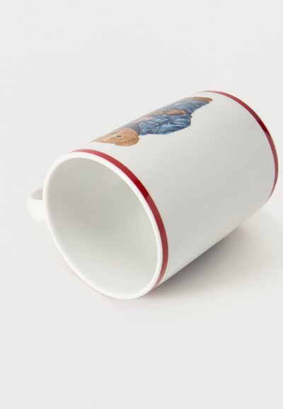 Cana ceramică albă cu margine și mâner roșu, având imprimat un ursuleț de pluș îmbrăcat într-o geacă albastră, întins pe o parte pe o suprafață albă.