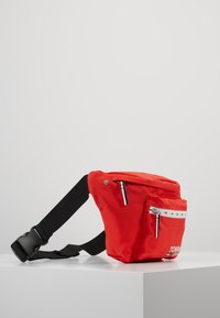 Rode stoffen heuptas met zwart verstelbaar riempje, voorzak met rits, klein logo en blauwe en zwarte ritslipjes op een witte ondergrond.