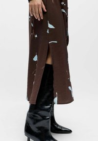 Mujer con un vestido marrón con estampado floral y una abertura lateral, y botas negras brillantes de tacón hasta la rodilla, posando frente a un fondo blanco liso.