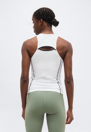 Γυναίκα με πλεγμένα μαλλιά, φορώντας λευκό αμάνικο τοπ racerback και πράσινα κολάν, όρθια με την πλάτη γυρισμένη σε απλό φόντο.