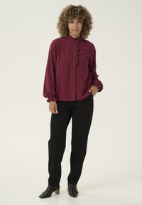 Burgundy button-up blouse with ruffled collar and cuffs, paired with black pants and heeled black ankle boots. Smooth texture, relaxed fit.