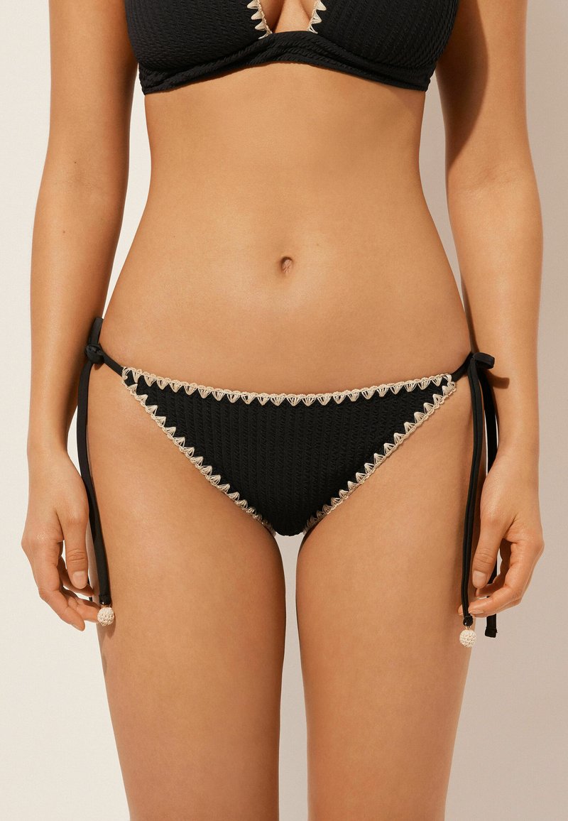 Czarne majtki bikini z teksturowanej tkaniny, w kształcie trójkąta, z kremowym koronkowym wykończeniem i regulowanymi wiązaniami po bokach.