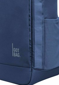 Námořnická modrá batoh s texturovanou tkaninou, šedé logo "GOT BAG" na boku, má hladký povrch a odolné popruhy. Kapsy viditelné na vnějšku.