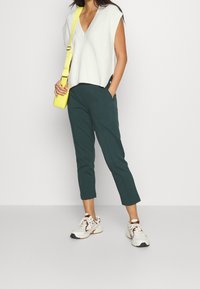 Top sin mangas de punto gris claro, pantalones de corte ajustado en color azul verdoso y zapatillas blancas con detalles en marrón; llevándose un pequeño bolso cruzado amarillo.