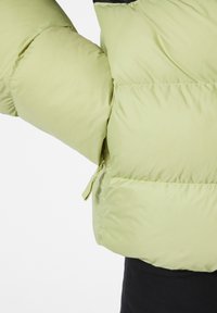 Chaqueta puff ligera de color verde claro con una textura acolchada, que presenta un detalle de cremallera en el lateral y acentos negros en los hombros.