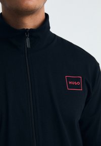Svart glidelåsjakke med høy krage, med en rød rektangulær logo med "HUGO" på venstre brystområde. Glatt tekstur, elastisk materiale.