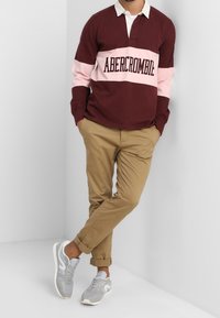 Bordeauxrood en roze polo met lange mouwen en witte kraag, met de tekst "ABERCROMBIE". Gecombineerd met beige chino broek en grijze sneakers.