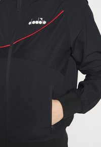 Svart zip-up-jacka med röd accentlinje, strukturerat tyg, elastiska ärmslut och sidofickor. Har en märkeslogotyp på bröstet.