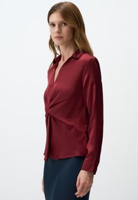 Red satin blouse with a deep V-neck and twisted front detail. Long sleeves with buttoned cuffs, paired with a dark blue skirt.