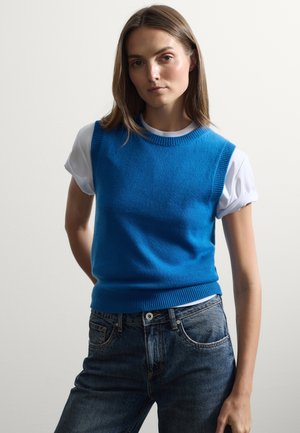 Blauwe gebreide mouwloze trui met geribde halslijn, gedragen over een witte T-shirt met opgerolde mouwen, gecombineerd met donkere spijkerbroeken. Soepele textuur.