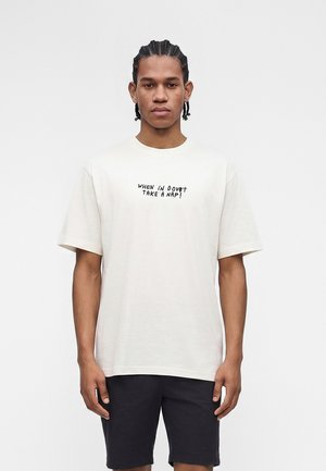 Νέος άνδρας με κοτσίδες, που φορά λευκό T-shirt με την επιγραφή "WHEN IN DOUBT TAKE A NAP!" και μαύρα σορτς, στέκεται μπροστά σε απλό φόντο.