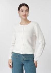 Νεαρή γυναίκα με σκούρα μαλλιά φοράει λευκή αφράτη ζακέτα με κουμπιά και μπλε τζιν, στέκεται μπροστά σε απλό φωτεινό φόντο.