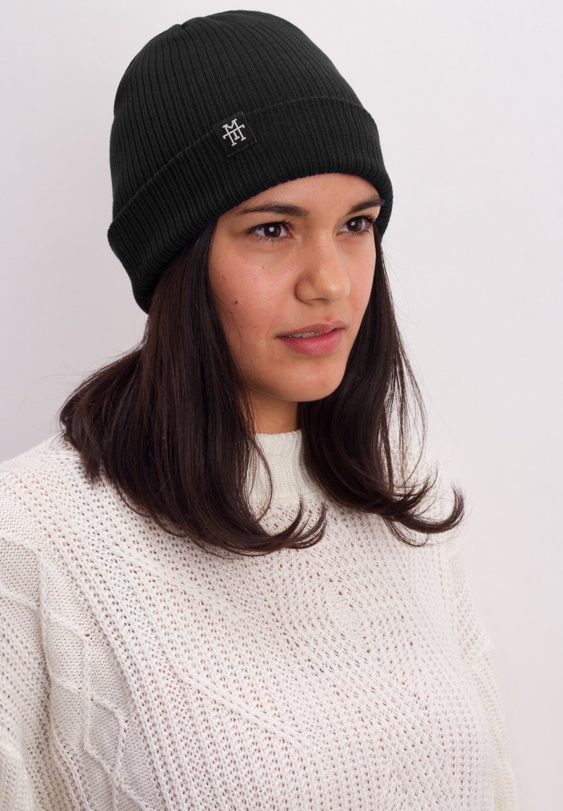 Black ribbed beanie with a logo patch. Soft, knit material, snug fit, with a fold-over cuff. Worn by a person with dark hair.