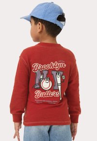 Rode lange mouw T-shirt met een grafische print van cartoon honkbal elementen en de tekst "Brooklyn Ballers" op de achterkant. Lichtblauwe pet.