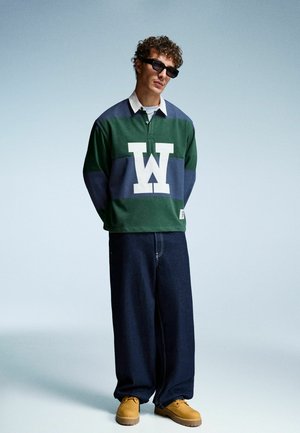Grön och marinrandig långärmad polo med vitt "W"-logotyp, ihop med vida mörk denimjeans och bruna arbetsskor.