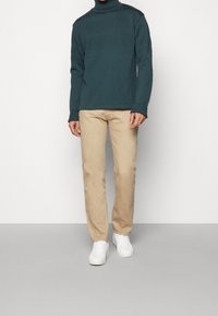 Turquoise knit turtleneck sweater with textured patterns paired with beige trousers and white sneakers. Slim fit; smooth fabric.