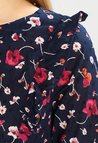 Granatowa tkanina w czerwony, biały i pomarańczowy wzór kwiatowy, z detalami falbanki w okolicy ramion.