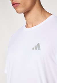 Camiseta deportiva blanca de tela transpirable, con cuello redondo y un detalle de logo plateado en la parte superior izquierda.
