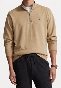 Sudadera color crema con media cremallera, mangas largas, puños acanalados y un pequeño logo en el pecho, combinada con pantalones deportivos oscuros.