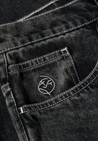 Un bolsillo de mezclilla negra con costuras blancas, que presenta un logo bordado. La textura desgastada resalta la apariencia usada de la tela.