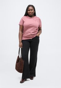 Donna in piedi con una mano nella tasca dei pantaloni neri, indossa una maglietta rosa con piccoli motivi a cuore, tiene una borsa marrone nell'altra mano.