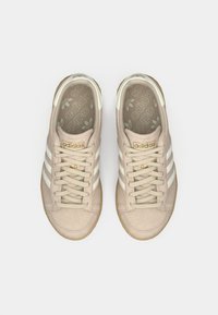 Svetlo bež Adidas športni copati z belimi črtami. Mehko semiš material, okrogla konica, vezanje spredaj in teksturiran gumijast podplat. Zlata logo podrobnost.