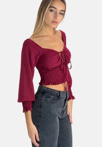 Bordeaux cropped blouse met een aangesloten, gerimpeld lijfje, klokmouwen en een striksluiting aan de voorkant, gecombineerd met high-waisted zwarte jeans.