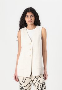 Beige linen vest with a straight cut, featuring a V-neck, five golden buttons, and two front pockets, layered over printed trousers.