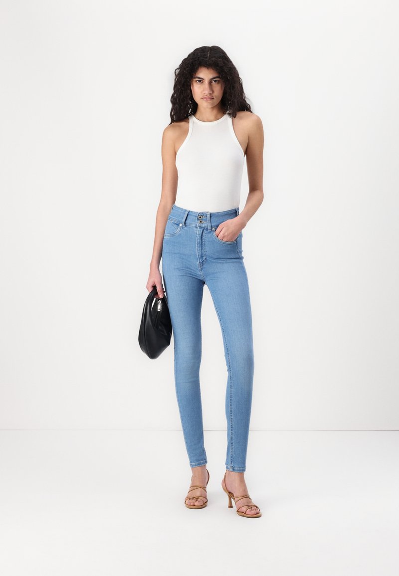 Hoch taillierte blue Jeans mit Slim Fit, heller Waschung, Zweiknopfverschluss und Gesäßtaschen. Kombiniert mit einem weißen Tanktop und einer schwarzen Handtasche.