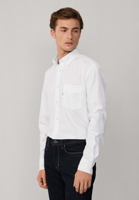 Camisa blanca de botones con un bolsillo en el pecho, mangas largas y cuello clásico; combinada con jeans oscuros que presentan detalles de costura visibles.