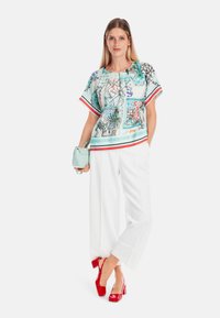 Mehrfarbige Seidenbluse mit kurzen Ärmeln, die mit abstrakten Mustern und Besatz versehen ist; kombiniert mit weißen, weit geschnittenen Hosen und roten Absätzen.