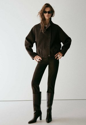 Femme portant une veste sombre surdimensionnée, un pantalon ajusté, des bottes à talons hautes jusqu'aux genoux et des lunettes de soleil, debout les mains sur les hanches devant un fond uni.