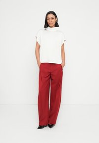 Valkoinen lyhythihainen paita korkealla kaula-aukolla, yhdistettynä punaisiin leveälahkeisiin housuihin ja mustiin teräväkärkisiin kenkiin. Yksinkertainen, selkeä muotoilu.