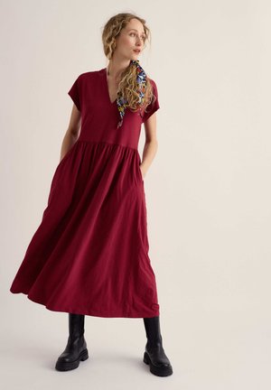 Femme portant une robe rouge ample à manches courtes, des bottes noires et un foulard coloré dans les cheveux, debout les mains dans les poches, regardant sur le côté.