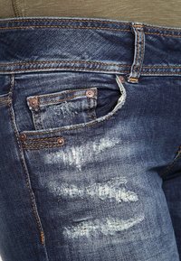 Jeans de mezclilla azul oscuro con textura desgastada, que presentan áreas deshilachadas, remaches de cobre y un diseño de pequeño bolsillo en la parte frontal.