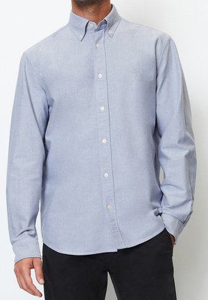 Hellblaues Hemd mit langen Ärmeln, getragen von einer Person mit einer Hand in der Tasche, kombiniert mit schwarzen Hosen.