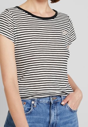 Woman wearing a black and white striped short-sleeve shirt tucked into blue denim jeans with one hand in pocket.